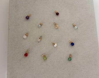 May include: A collection of eleven small jewelry charms, each featuring a single round gemstone bead in various colors including purple, red, blue, green, and white, accented with a small gold-toned metal jump ring at the top for attachment.