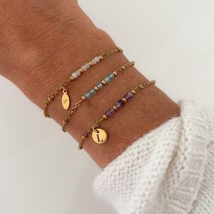 Könnte beinhalten: Drei goldfarbene Kettenarmbänder mit kleinen Perlen und Anhängern. Das oberste Armband hat weiße Perlen und einen ovalen Anhänger mit dem Buchstaben S. Das mittlere Armband hat blaue Perlen. Das untere Armband hat violette Perlen und einen runden Anhänger mit dem Buchstaben L.