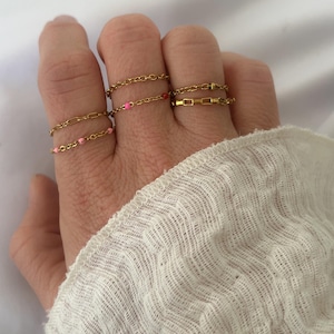 Pode incluir: Uma mão usando cinco anéis de corrente de ouro com detalhes rosa e vermelho. Os anéis estão empilhados em dois dedos.