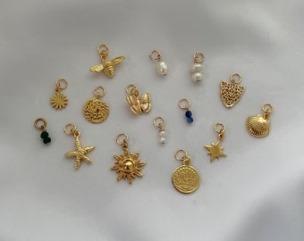 May include: A collection of 14 gold-toned charms, including a bee, butterfly, sun, star, shell, and coin. The charms are arranged on a white surface.