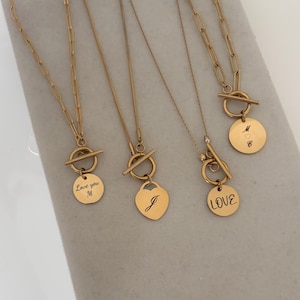 Puede incluir: Cuatro collares dorados con cierres de barra y colgantes grabados. Los colgantes incluyen formas circulares y de corazón con inscripciones como Love you M, J, LOVE y M con un símbolo de corazón. Las cadenas varían en estilo de eslabón.