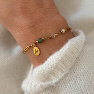 Può includere: Bracciale dorato con design a catena e un piccolo ciondolo ovale con la lettera "M". Il bracciale include perline verdi, bianche e trasparenti. Il bracciale è indossato su un polso.