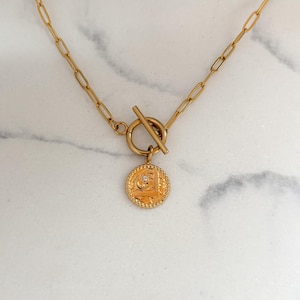 Puede incluir: Collar dorado con un colgante de moneda con un pequeño diamante. El collar tiene un cierre de palanca y un diseño de cadena de eslabones. El colgante de moneda tiene un diseño intrincado.