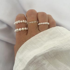 Könnte beinhalten: Drei Perlenringe an einer Hand. Die Ringe bestehen aus kleinen weißen Perlen und sind übereinander gestapelt.