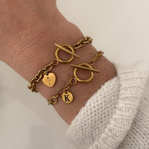 Peut inclure: Deux bracelets dorés avec fermoirs à bascule. L'un des bracelets comporte un charm en forme de cœur avec les lettres "A L" et une petite flèche. L'autre bracelet a un charm circulaire avec la lettre "K". Les bracelets sont portés au poignet.