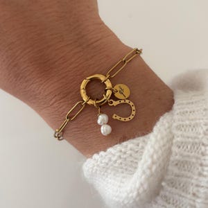 Könnte beinhalten: Ein goldfarbenes Gliederarmband mit einem runden Verschluss. Am Verschluss befinden sich drei Anhänger: eine kleine runde Scheibe mit der Gravur JL, ein Hufeisenanhänger und ein Paar kleiner, unregelmäßiger weißer Süßwasserperlen.