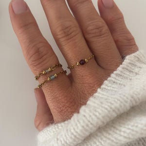 Könnte beinhalten: Drei zarte goldfarbene Kettenringe mit kleinen Metallperlen und zentralen Steinen. Zwei Ringe werden am Zeigefinger getragen, jeder mit einem grauen Stein, und ein Ring wird am Mittelfinger getragen, der einen einzelnen dunkelroten Stein aufweist.
