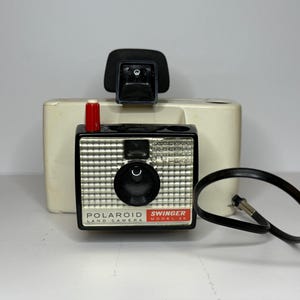 May include: A vintage Polaroid Swinger Model 20 land camera. The camera is black and silver with a large lens and a red button. The camera has a black strap and is set against a white background.