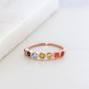 May include: A rose gold band ring with seven colorful gemstones. The gemstones are arranged in a rainbow pattern, starting with purple and ending with red.