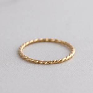May include: A delicate gold ring with a twisted rope design. The ring is thin and has a polished finish, reflecting light. The jewelry is set against a neutral background, highlighting its simple elegance.