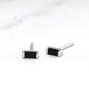 Peut inclure: Une paire de petites boucles d'oreilles à clous rectangulaires. Chaque boucle d'oreille présente une pierre noire rectangulaire sertie dans un métal argenté. Les boucles d'oreilles sont présentées sur une surface blanche.