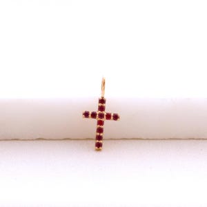 May include: A gold-colored cross pendant adorned with small, vibrant red gemstones. The cross features a loop at the top for a chain and is set against a soft, white background. The gemstones are evenly spaced, creating a symmetrical design.