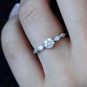 May include: A silver ring with a large, round, clear gemstone at the center, flanked by smaller, marquise-shaped stones. The band features delicate detailing. The ring is shown on a person's finger.