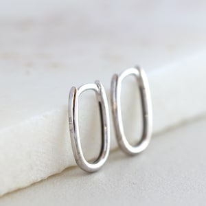 May include: A pair of oval-shaped silver earrings. The earrings have a simple, minimalist design with a smooth, polished finish. The text "JL 10K" is stamped on the side of one earring. The earrings are displayed on a white surface.