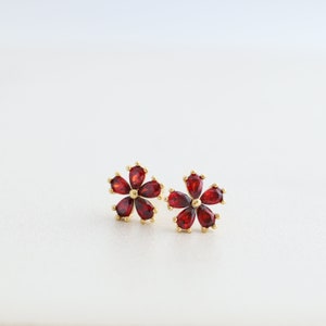 May include: A pair of gold-colored flower stud earrings. Each earring features five teardrop-shaped, deep red gemstones arranged to resemble a flower. The earrings are set against a white background.