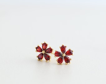 May include: A pair of gold-colored flower stud earrings. Each earring features five teardrop-shaped, deep red gemstones arranged to resemble a flower. The earrings are set against a white background.