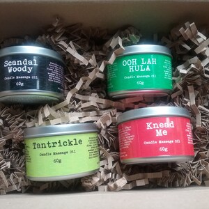 Puede incluir: Cuatro peque&ntilde;as latas de metal con etiquetas que contienen aceite de masaje para velas. Las latas est&aacute;n en una caja de cart&oacute;n marr&oacute;n llena de papel de embalaje marr&oacute;n. Las etiquetas son negras, verdes, amarillas y rojas. Las etiquetas dicen "Scandal Woody", "Ooh Lah Hula", "Tantrickle" y "Knead Me".