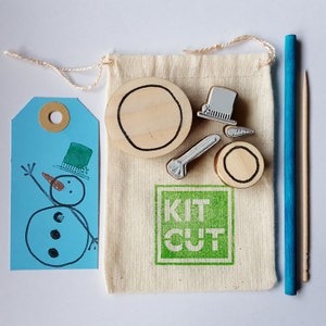 May include: A kit for making rubber stamps with a snowman, hat, and other winter-themed designs. The kit includes a small bag, wooden blocks, and a blue and white stamp. The bag has the text "Kit Cut" printed on it.