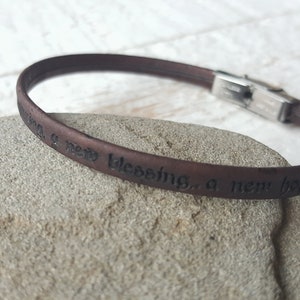 May include: A brown leather bracelet with a silver clasp. The bracelet has the words "a new blessing, a new hope" engraved on it.