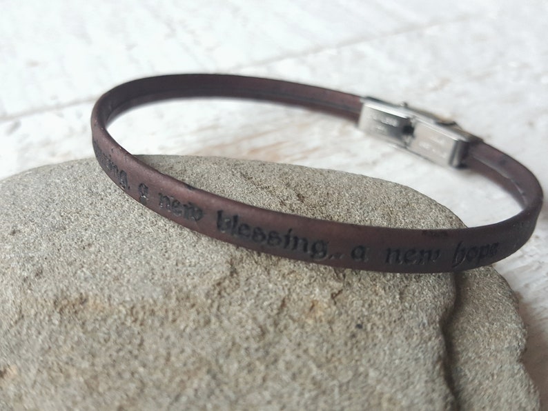 May include: A brown leather bracelet with a silver clasp. The bracelet has the words "a new blessing, a new hope" engraved on it.