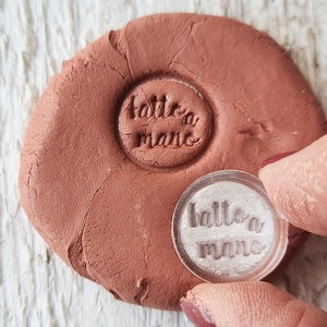 Könnte beinhalten: Ein runder, transparenter Stempel mit dem Text "fatto a mano" in einer kursivschriftlichen Schriftart. Der Stempel wird verwendet, um einen Abdruck in einem Stück braunem Ton zu erstellen.