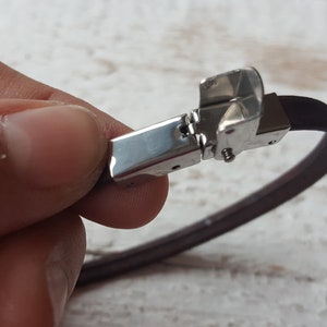 May include: A brown leather bracelet with a silver clasp. The clasp is open and the bracelet is being held by a hand.