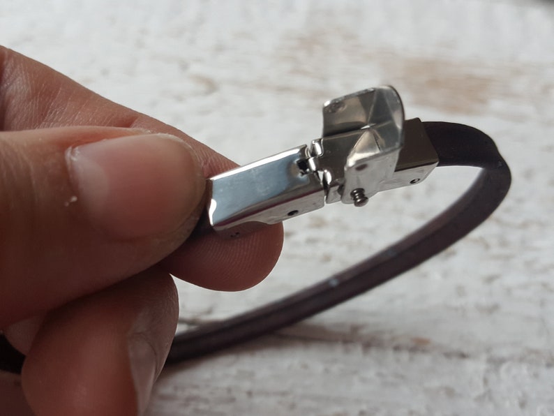 May include: A brown leather bracelet with a silver clasp. The clasp is open and the bracelet is being held by a hand.