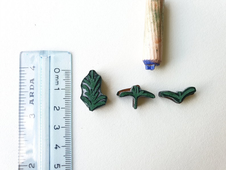 May include: Four green rubber stamps with leaf designs. The stamps are approximately 1 cm in size. A wooden roller stamp is also visible in the image.