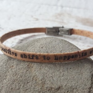 May include: A cork bracelet with a silver clasp and the inscription "a positive shift to happen is over!"