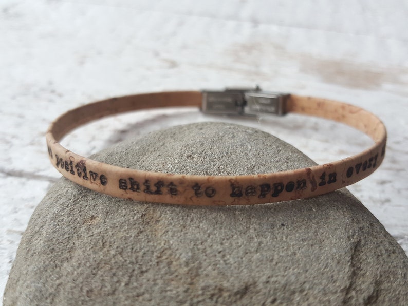 May include: A cork bracelet with a silver clasp and the inscription "a positive shift to happen is over!"
