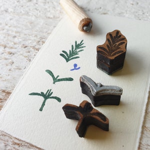 May include: A set of four wooden stamps featuring various leaf designs. The stamps are displayed on a white card with green and blue ink impressions. These stamps are ideal for crafting and scrapbooking.
