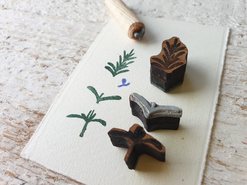 May include: A set of four wooden stamps featuring various leaf designs. The stamps are displayed on a white card with green and blue ink impressions. These stamps are ideal for crafting and scrapbooking.