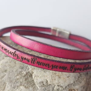 May include: A pink leather wrap bracelet with a silver clasp. The bracelet has the inscription "Believe in miracles, you'll never see one. If you believe in m"