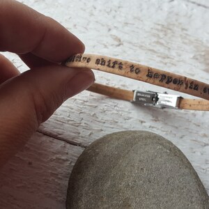 May include: Cork bracelet with a stainless steel clasp. The bracelet has the words "a positive shift to happen in every day" engraved on it.