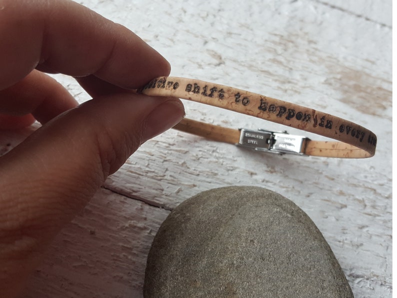 May include: Cork bracelet with a stainless steel clasp. The bracelet has the words "a positive shift to happen in every day" engraved on it.
