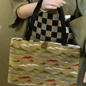May include: A beige tote bag with a black handle and a button closure. The bag has a pattern of green, blue, and red mountains and white clouds.