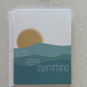 May include: Greeting card with a minimalist design featuring a golden sun setting over teal waves. The words "just keep swimming" are printed in a clean, sans-serif font. The card is white with a matching envelope.