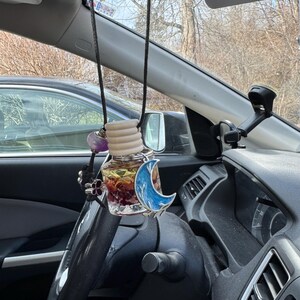 Puede incluir: Un ambientador para coche colgado del espejo retrovisor. El ambientador es una pequeña botella de vidrio llena de flores secas y un amuleto de media luna azul. La botella está unida a un cordón negro con una cuenta de cristal morado.