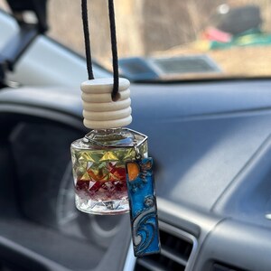 Puede incluir: Una botella de vidrio transparente con una tapa de madera y un amuleto colorido colgando de ella. La botella está llena de un líquido y pequeñas flores. El amuleto es una pieza rectangular de metal con un diseño azul, naranja y amarillo.