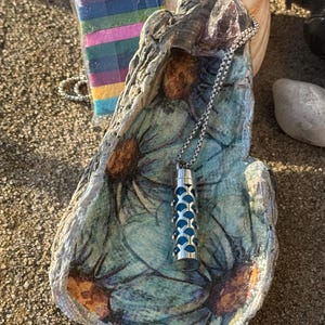 May include: A silver-colored pendant necklace with a blue interior, displayed on a painted seashell. The seashell is teal with floral accents. A small packet of colorful felt squares is visible in the background.