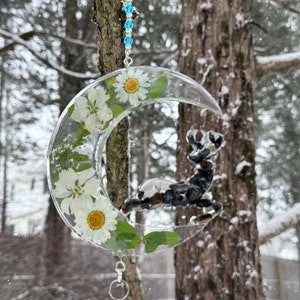 May include: A crescent moon-shaped suncatcher with white daisies and green leaves encased in clear resin. A small, dark-colored deer figure is inside the moon. The suncatcher hangs from a string of blue beads and a silver chain.