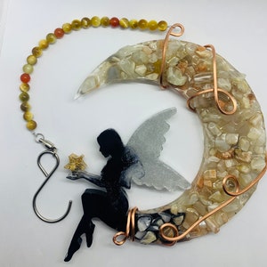 May include: A black fairy silhouette holding a gold star sits on a crescent moon made of clear resin with embedded white and brown stones. The moon is wrapped with copper wire and hangs from a beaded chain.