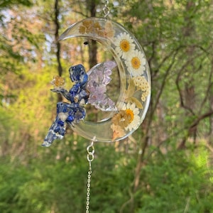May include: A crescent moon-shaped suncatcher with a fairy figurine. The moon is filled with dried daisies and other flowers. The fairy is blue and purple, holding a golden object. A silver chain is attached for hanging.
