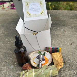May include: A white box with a floral design on the lid, containing a necklace, essential oil bottle, crystals, a seashell, a bundle of sage, and a piece of wood. The necklace has a silver chain and a rectangular pendant with the number 3.