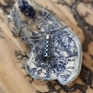 May include: A decorative oyster shell-shaped dish with a blue and white floral pattern. A silver necklace and a silver pendant with blue accents rest inside the dish. The dish sits on a wooden surface.