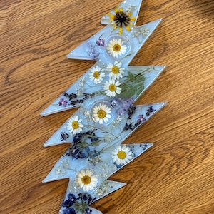 May include: A light blue resin lightning bolt decoration with embedded dried flowers. The bolt is filled with daisies, purple flowers, and a yellow sunflower. The decoration is approximately 12 inches long.