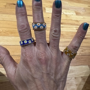 May include: A hand wearing three beaded rings. The rings are made of small, colorful beads in a floral pattern. The rings are worn on the index, middle, and ring fingers.