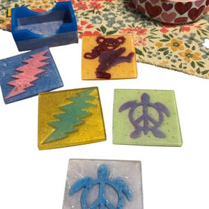 Könnte beinhalten: Eine Sammlung farbenfroher Harzuntersetzer mit verschiedenen Designs. Die Untersetzer zeigen einen Blitz, einen tanzenden Bären, eine Peace-Zeichen-Schildkröte und andere einzigartige Grafiken. Die Untersetzer sind quadratisch und haben eine glänzende Oberfläche, ideal für die Inneneinrichtung.
