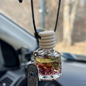 Puede incluir: Una botella de vidrio transparente llena de líquido amarillo y cristales rosas, colgando de una cuerda negra. La botella tiene un tapón de madera y un amuleto plateado con un ojo azul y un diseño de mano.