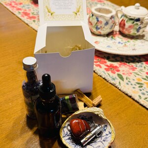 May include: A white box with text, a small glass bottle, a brown dropper bottle, a seashell with a red heart-shaped stone, a bundle of red sage, and a small bundle of wood sticks. The box contains a list of items. The items are arranged on a wooden table.